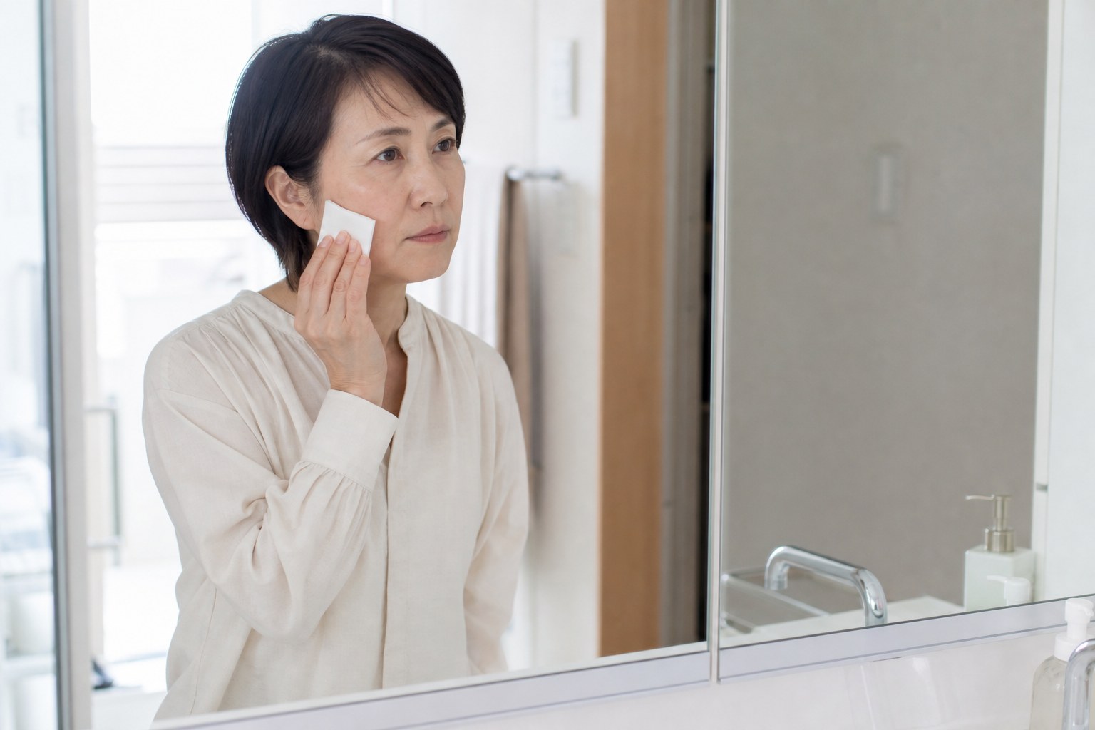 洗面台の鏡の前でコットンを頬に当てようとして手を止める50代女性